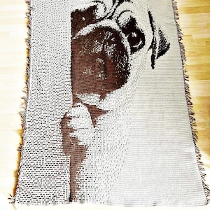 Op de afbeelding: Een geweven sprei met een gedetailleerde afbeelding van het gezicht van een mopshond. De hond is bruin op een lichtgrijze achtergrond. De deken heeft een gefranjerde rand.