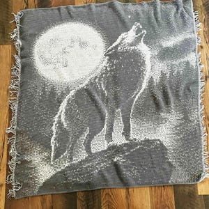 May include: A gray and white throw blanket featuring a howling wolf silhouette against a full moon. The blanket has a fringed edge and a textured design, perfect for adding a touch of nature-inspired decor to any space.