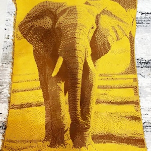 Puede incluir: Una manta de ganchillo amarilla y marrón con una imagen detallada de un elefante. El elefante está representado en un estilo pixelado, con líneas horizontales en el fondo. La manta tiene un borde con flecos.