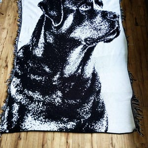 Puede incluir: Una manta tejida en blanco y negro con un retrato detallado de un perro. La cabeza y los hombros del perro se representan en un estilo pixelado de alto contraste. La manta tiene un borde con flecos.