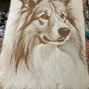 Puede incluir: Una manta tejida con un retrato detallado de un perro. La imagen del perro está en tonos marrones sobre un fondo crema. La manta tiene un borde con flecos.