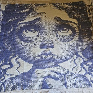 Puede incluir: Una manta tejida con un retrato detallado de un rostro en azul y blanco. El diseño está pixelado, creando una textura única. La manta tiene un borde con flecos.