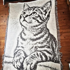 Puede incluir: Una manta tejida con una imagen detallada de un gato. El gato está representado en tonos grises y negros sobre un fondo crema. La manta tiene un borde con flecos y está colocada sobre un suelo de madera.