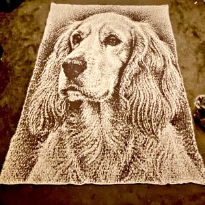 Op de afbeelding: Een grote, rechthoekige deken met een gedetailleerd portret van een hondengezicht. De vacht van de hond is afgebeeld in een getextureerde, lichtbeige kleur tegen een donkerbruine achtergrond. De deken is waarschijnlijk gemaakt van een zacht materiaal.