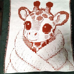 Puede incluir: Una manta de ganchillo rectangular con un diseño bicolor en blanco y naranja quemado. El patrón representa una cabeza de jirafa estilizada con ojos grandes, orejas y osiconos, renderizada en un estilo pixelado sobre un fondo blanco sólido.