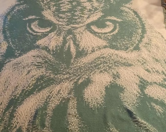 May include: A large, rectangular blanket featuring a detailed owl design. The owl's face is prominently displayed in shades of green and white, with fringed edges. The blanket is made of a soft, woven material.