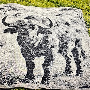 May include: A rectangular woven blanket featuring a high-contrast black and white graphic design of a Cape buffalo. The animal is depicted in a standing position, showcasing its large curved horns and textured coat against a light background.