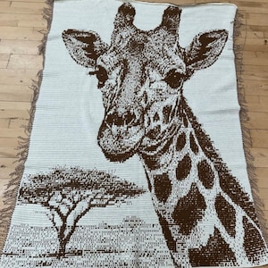 Könnte beinhalten: Eine gewebte Decke mit einem braunen Giraffenmuster auf hellem Beige. Der Kopf und Hals der Giraffe stehen im Mittelpunkt, mit einer Baumsilhouette unten links. Die Decke hat einen Fransenrand.