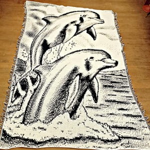 Puede incluir: Una manta tejida de color crema con una ilustración en blanco y negro de dos delfines saltando del agua. La manta tiene un borde con flecos y es de forma rectangular. Los delfines son el punto focal del diseño.