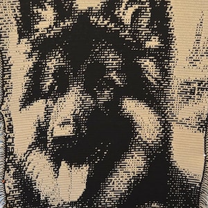 Puede incluir: Una gran manta de ganchillo hecha a mano con una imagen detallada en negro y beige de la cara de un perro. La lengua del perro es visible. La manta tiene un borde con flecos y está hecha con un diseño pixelado.