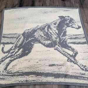 Puede incluir: Una manta rectangular de ganchillo que presenta la silueta de un galgo gris corriendo, centrada sobre un fondo de color crema. La manta tiene un borde gris sólido y una apariencia tejida y texturizada en todo el diseño.