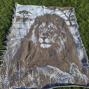Puede incluir: Una manta tejida marrón y blanca con una imagen detallada de la cara de un león y un paisaje de sabana. La manta tiene un borde con flecos y se muestra sobre hierba verde.