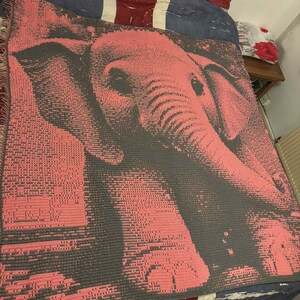 May include: A pink and gray crocheted blanket featuring an elephant design. The blanket is draped over a bed with a Union Jack-themed pillow and bedding. The elephant's features are detailed in the crochet work.