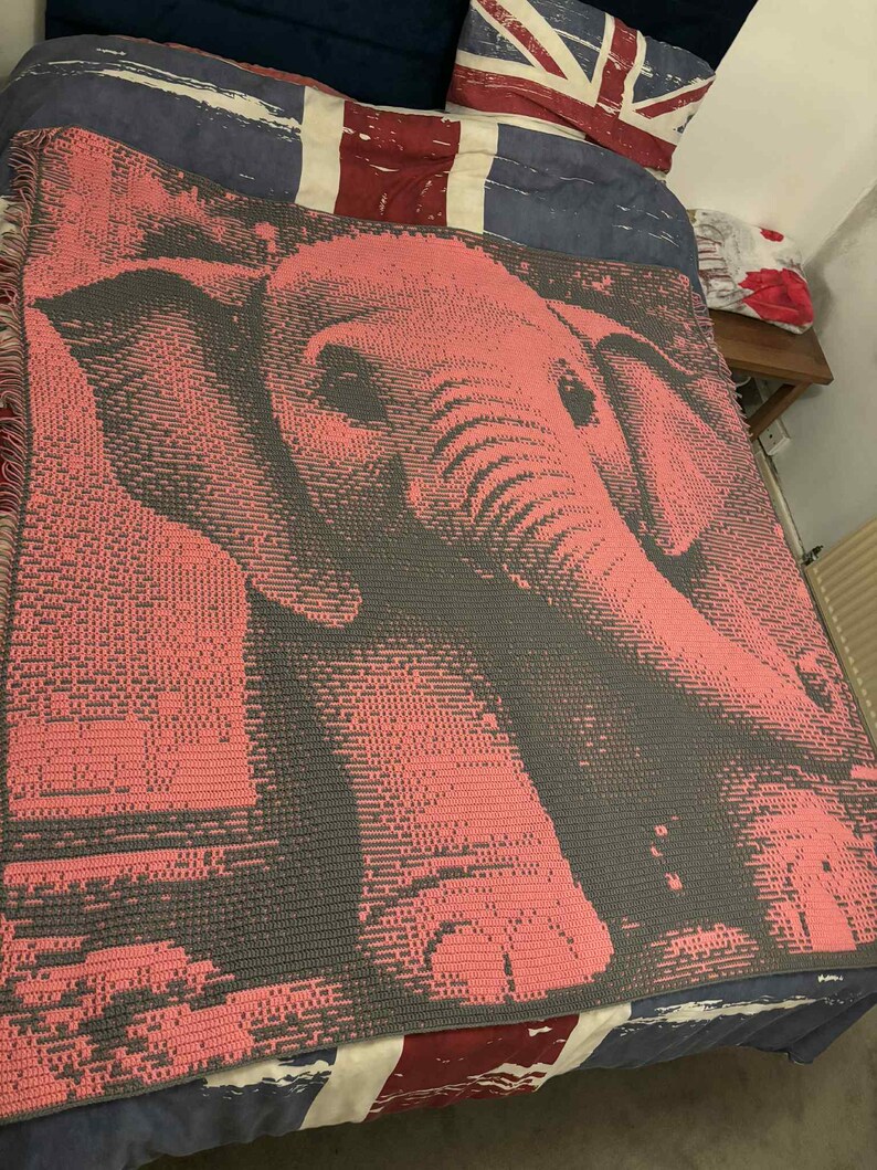 May include: A pink and gray crocheted blanket featuring an elephant design. The blanket is draped over a bed with a Union Jack-themed pillow and bedding. The elephant's features are detailed in the crochet work.