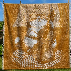 Puede incluir: Una manta tejida de color amarillo mostaza y blanco con un gráfico detallado de un gato tocando el contrabajo. La manta presenta una textura de rejilla en toda su superficie y está rematada con flecos blancos en los dos bordes laterales.