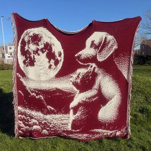 Puede incluir: Una manta burdeos con un gráfico blanco que representa a un perro mirando una luna llena. La manta tiene un borde con flecos y se muestra al aire libre en un día soleado. El diseño del perro y la luna contrasta con el fondo burdeos.