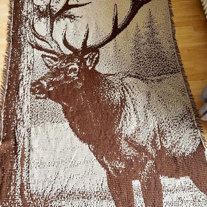 May include: A brown and beige woven throw blanket featuring a detailed image of a stag with large antlers. The blanket has a fringed edge and a forest scene background. The stag is the focal point, rendered in a pixelated style.