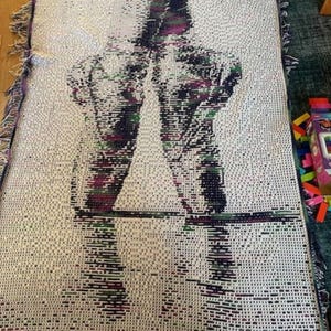 Puede incluir: Una manta tejida con un diseño abstracto de bailarinas de ballet con zapatillas de punta. La manta tiene un fondo blanco con un patrón de negro, morado y verde. Los bordes están acabados con flecos.