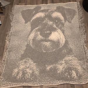 May include: A gray and white woven throw blanket featuring a detailed portrait of a Schnauzer dog. The blanket has a fringed edge and a textured appearance, suitable for home decor.