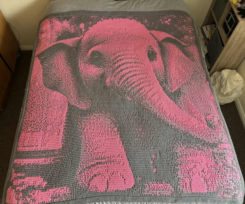 May include: A handmade blanket featuring a pixelated image of an elephant in shades of pink and gray. The blanket is spread across a bed, showcasing the detailed design and craftsmanship. The elephant's form is clearly defined against the gray background.
