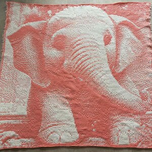 May include: Coral and white crocheted blanket featuring a detailed elephant design. The blanket showcases the elephant's head and trunk, with intricate stitchwork creating a textured appearance. The blanket is square-shaped and has a fringed edge.