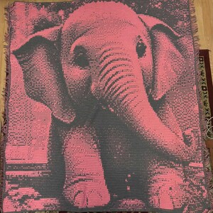 May include: Pink and gray elephant throw blanket. The blanket features a detailed pixelated design of an elephant's face and body. The blanket has a fringed edge and is made of a soft material. The color palette is primarily pink and gray.