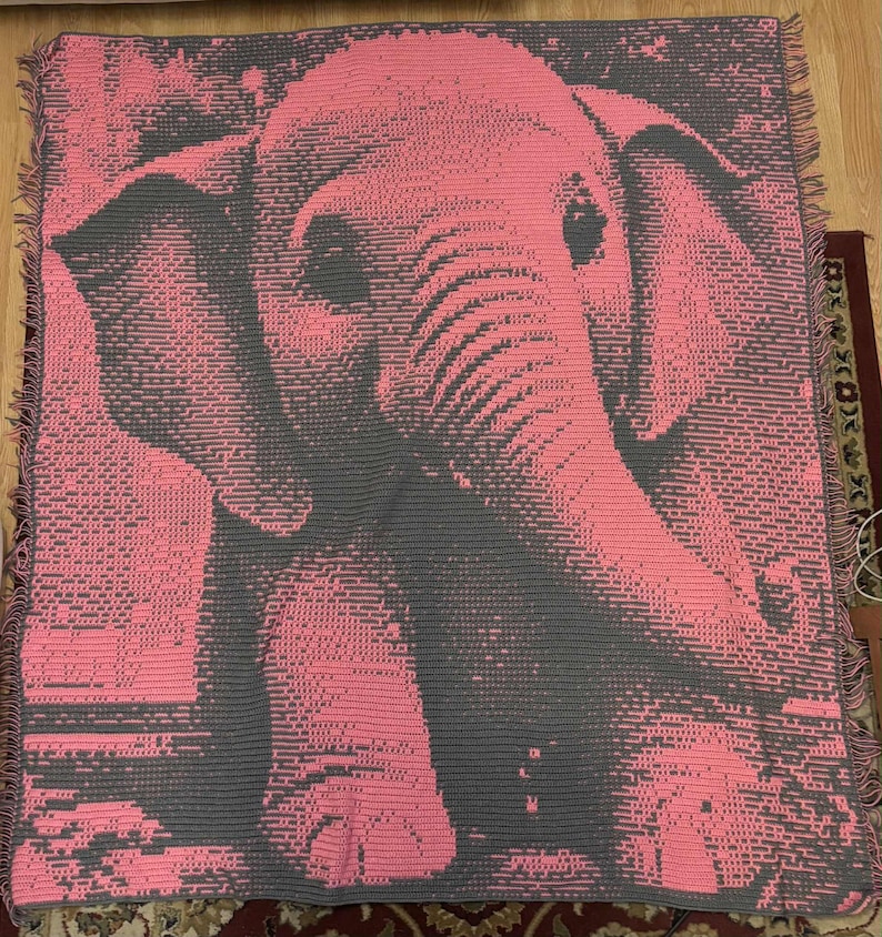 May include: Pink and gray elephant throw blanket. The blanket features a detailed pixelated design of an elephant's face and body. The blanket has a fringed edge and is made of a soft material. The color palette is primarily pink and gray.