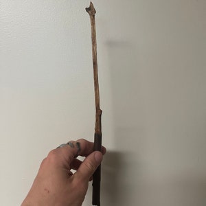 Puede incluir: Una varita de madera hecha a mano con una punta. La varita está hecha de una rama natural, con un mango marrón oscuro que hace la transición a un eje marrón claro. La varita se sostiene contra una pared blanca lisa.