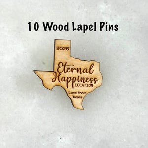 May include: Ten wood lapel pins. Each pin is shaped like the state of Texas, with the words "Eternal Happiness LOCATION" and "Love From Texas" engraved. The year "2026" is also engraved on the pin.