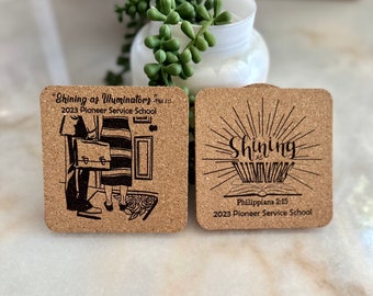 May include: Two cork coasters with black text and images. The first coaster features a black and white illustration of two people holding briefcases and standing in a doorway. The text reads "Shining as Illuminators" and "2023 Pioneer Service School". The second coaster features a black and white illustration of an open book with rays of light emanating from it. The text reads "Shining as Illuminators", "Philippians 2:15", and "2023 Pioneer Service School".