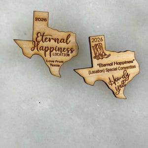 May include: Two wooden Texas-shaped ornaments. One reads "Eternal Happiness" with "Love From Texas" and the year "2026". The other says "Eternal Happiness" with "Howdy Y'all" and the year "2026".