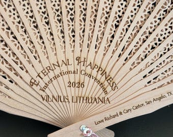Puede incluir: Un abanico plegable de madera clara con intrincados diseños calados. El abanico tiene el texto "Eternal Happiness International Convention 2026 Vilnius, Lituania". Una borla rosa cuelga de la base.