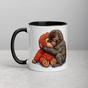 May include: White ceramic mug with a black handle and rim. The mug features a colorful illustration of a brown monkey hugging a red stuffed animal monkey. The design is printed on the front of the mug.