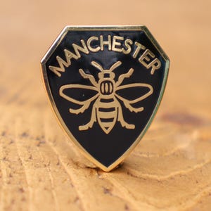 May include: A black and gold enamel pin in a shield shape. The pin features a gold bee design and the word "MANCHESTER" in gold lettering. The pin has a gold border. The pin is set against a wooden surface.