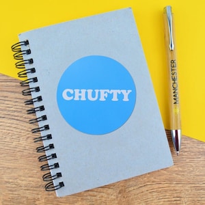 May include: A spiral-bound notebook with a blue circle and the word "CHUFTY" in white. A pen with "MANCHESTER" printed on it rests beside the notebook. The background is yellow and the surface is wood.