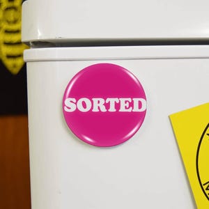 May include: A circular, magenta fridge magnet displaying the word "SORTED" in white, bold lettering. The magnet is attached to a white surface. Other decorative items are visible in the background.