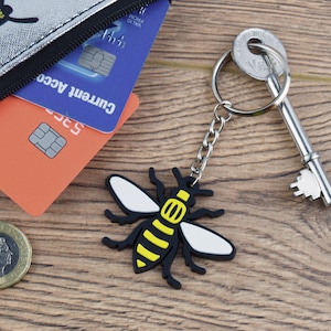 Peut inclure: Un porte-clés en forme d'abeille noir et jaune avec des ailes blanches. Le porte-clés est attaché à un anneau de clé argenté et à une clé argentée.
