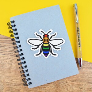 May include: A light blue spiral notebook with a colorful bee sticker and a pen. The bee sticker has a rainbow design on its body and white wings. The pen is bamboo-colored with the word "MANCHESTER" printed on it.