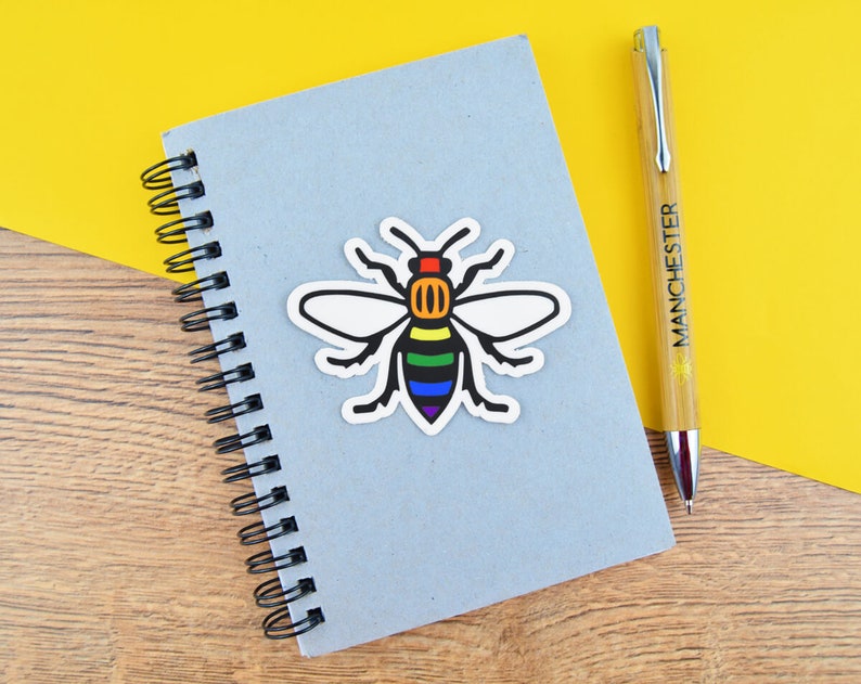 May include: A light blue spiral notebook with a colorful bee sticker and a pen. The bee sticker has a rainbow design on its body and white wings. The pen is bamboo-colored with the word "MANCHESTER" printed on it.