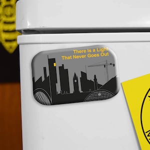 May include: A rectangular magnet with a gray background featuring a black silhouette of a city skyline. The text "There Is a Light That Never Goes Out" is printed on the magnet. A yellow sticker with the word "MAN" is visible.