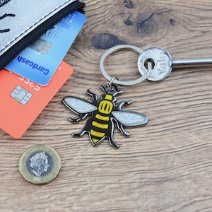 Puede incluir: Un llavero plateado con un dije de abeja negro y amarillo. La abeja tiene brillo en sus alas. El llavero está unido a un anillo de llave plateado y una llave plateada con la palabra "Yale" en ella.