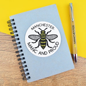 May include: A grey spiral-bound notebook with a round sticker showing a black and yellow bee and the words "MANCHESTER. MANC AND PROUD." A pen with "MANCHESTER" printed on it lies next to the notebook on a yellow surface.