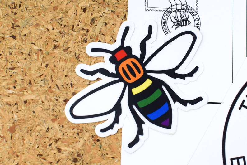 May include: A sticker featuring a stylized bee with a rainbow-colored body and black wings. The bee is set against a white background, with a corkboard in the background. The sticker has a white border.