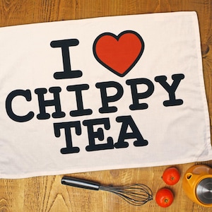 May include: A white tea towel with the words "I ❤️ CHIPPY TEA" in black, with a red heart. The towel is on a wooden surface, with a whisk, two tomatoes, and a yellow kitchen gadget.