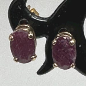 May include: A pair of oval-shaped ruby earrings set in gold. The earrings are a deep red color and have a polished finish.