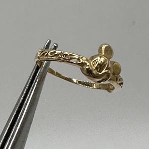 May include: A gold-toned ring featuring a Mickey Mouse silhouette. The ring has a delicate, ornate design.