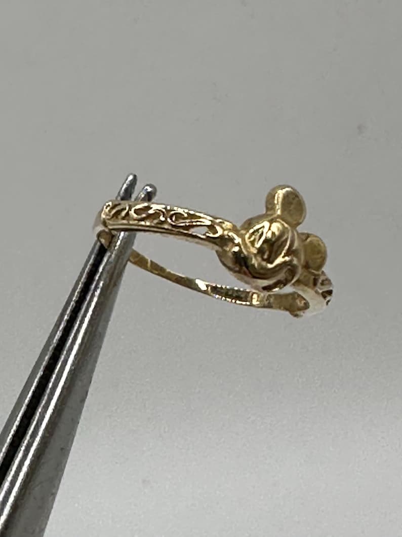 May include: A gold-toned ring featuring a Mickey Mouse silhouette. The ring has a delicate, ornate design.