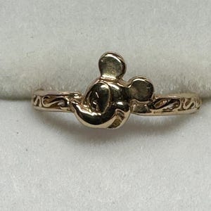 May include: A gold-toned ring featuring a Mickey Mouse head design. The ring band is thin and has a delicate, swirling pattern.