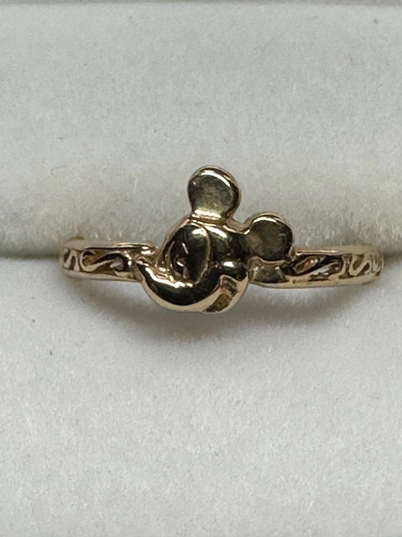 May include: A gold-toned ring featuring a Mickey Mouse head design. The ring band is thin and has a delicate, swirling pattern.