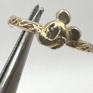 May include: A gold-colored ring with a Mickey Mouse head design. The ring is detailed with a floral pattern.